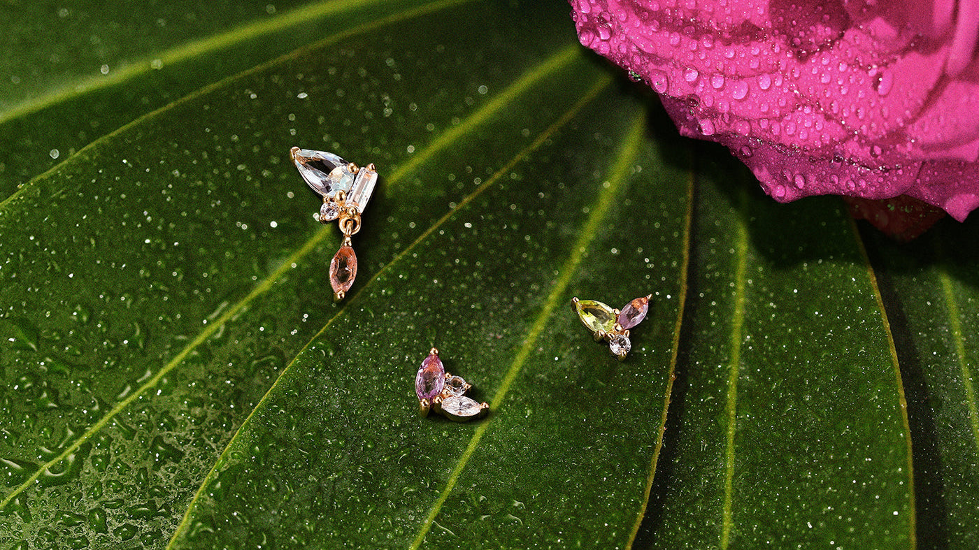 Delicate gold stud earrings featuring pastel pink and clear gemstones resting on a wet green leaf near a pink flower.