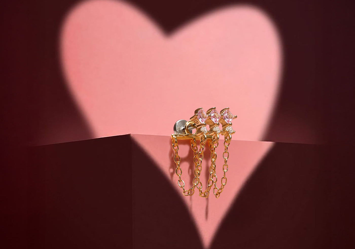 Gold piercing stud with square pink gems and dangling chains, displayed against a pink heart shadow background.