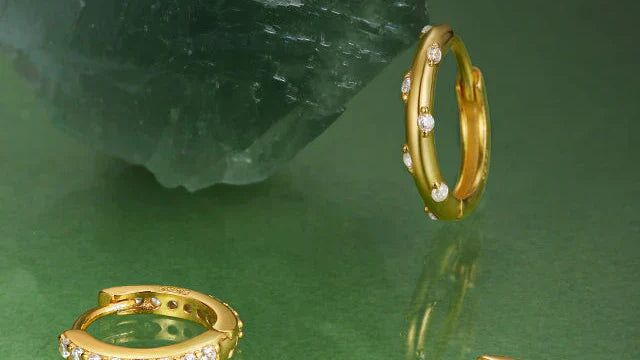 Small gold huggie hoop earrings embedded with clear gemstones arranged on a green surface near a raw crystal rock.