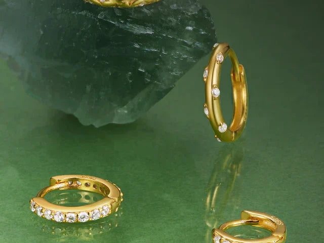 Small gold huggie hoop earrings embedded with clear gemstones arranged on a green surface near a raw crystal rock.