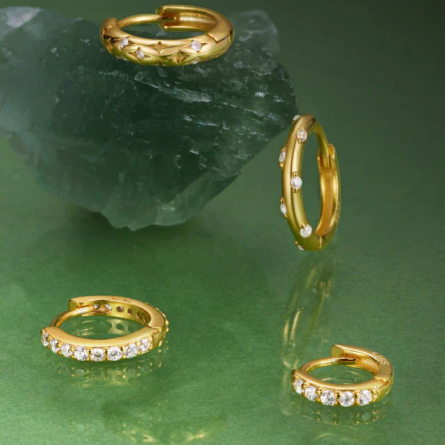 Small gold huggie hoop earrings embedded with clear gemstones arranged on a green surface near a raw crystal rock.