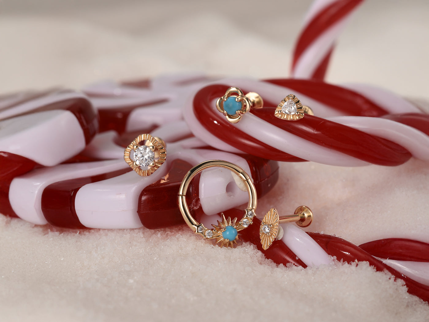 A festive holiday scene featuring Kosiner's gold and turquoise piercing jewelry, including studs and a hoop, artfully arranged on a candy cane.