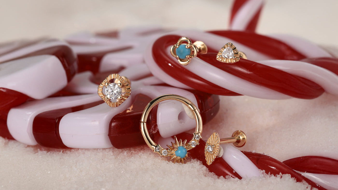 A festive holiday scene featuring Kosiner's gold and turquoise piercing jewelry, including studs and a hoop, artfully arranged on a candy cane.