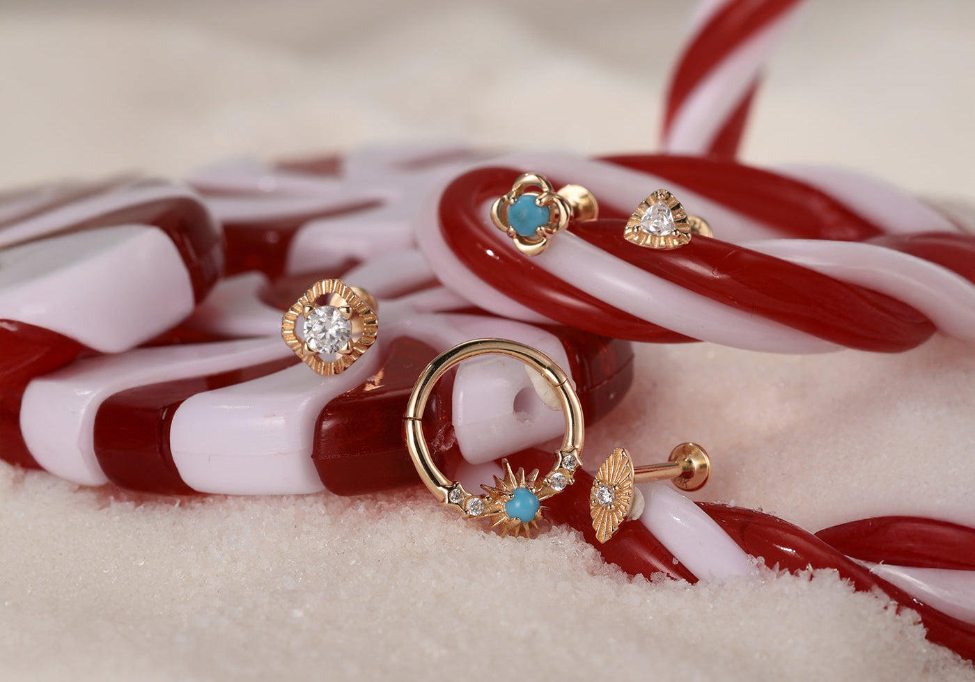 A festive holiday scene featuring Kosiner's gold and turquoise piercing jewelry, including studs and a hoop, artfully arranged on a candy cane.