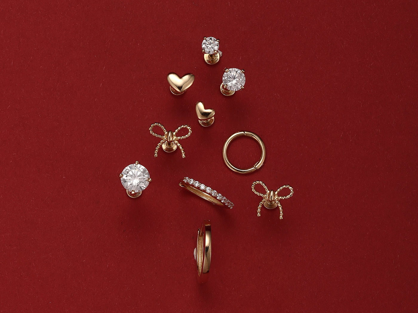 An assortment of classic gold hypoallergenic earrings, including studs and hoops, arranged in a Christmas tree shape on a festive red background.