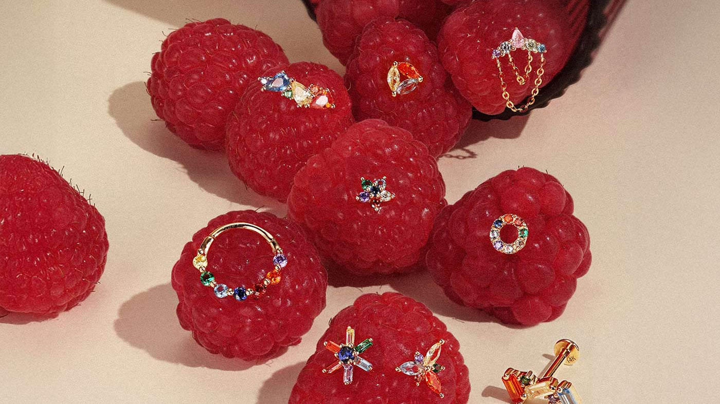 Assorted colorful rainbow gold earrings including clicker hoops and studs displayed on fresh raspberries for ear stacking ideas.