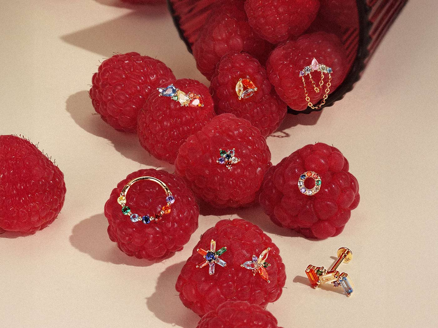 Assorted colorful rainbow gold earrings including clicker hoops and studs displayed on fresh raspberries for ear stacking ideas.