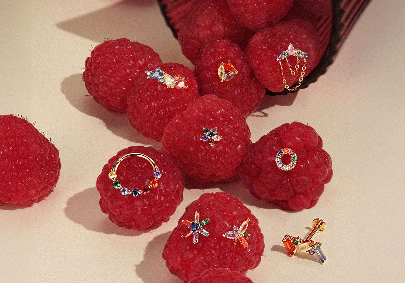 Assorted colorful rainbow gold earrings including clicker hoops and studs displayed on fresh raspberries for ear stacking ideas.