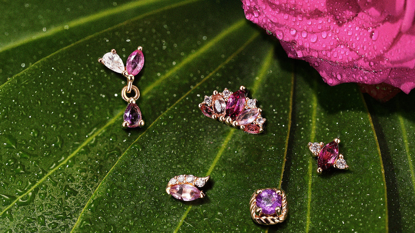 Collection of hypoallergenic titanium piercing studs featuring vibrant purple gemstones resting on a natural green leaf with water droplets.