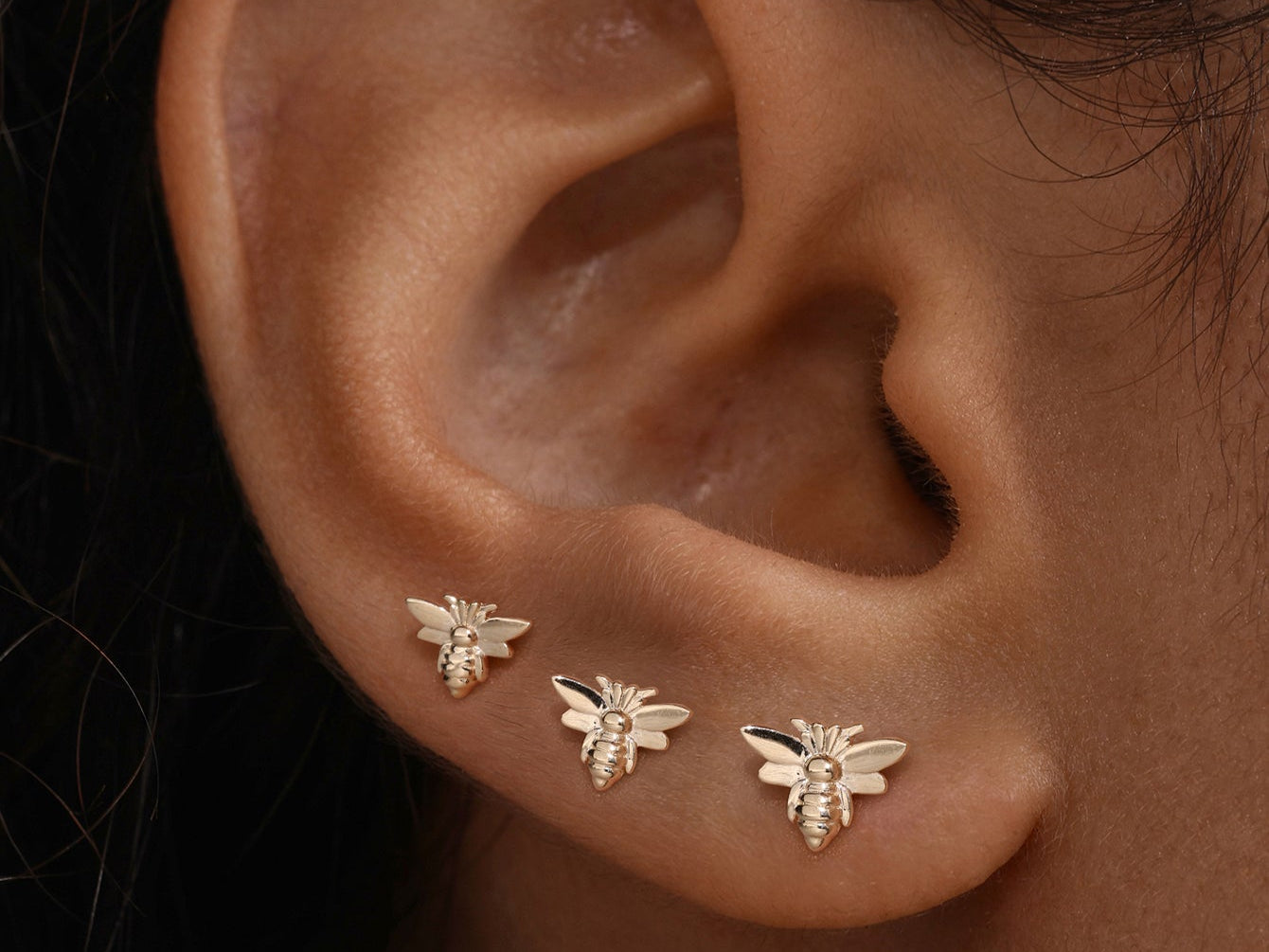 Close-up of three gold bee-shaped stud earrings on a healed earlobe, representing safe jewelry for piercing aftercare.
