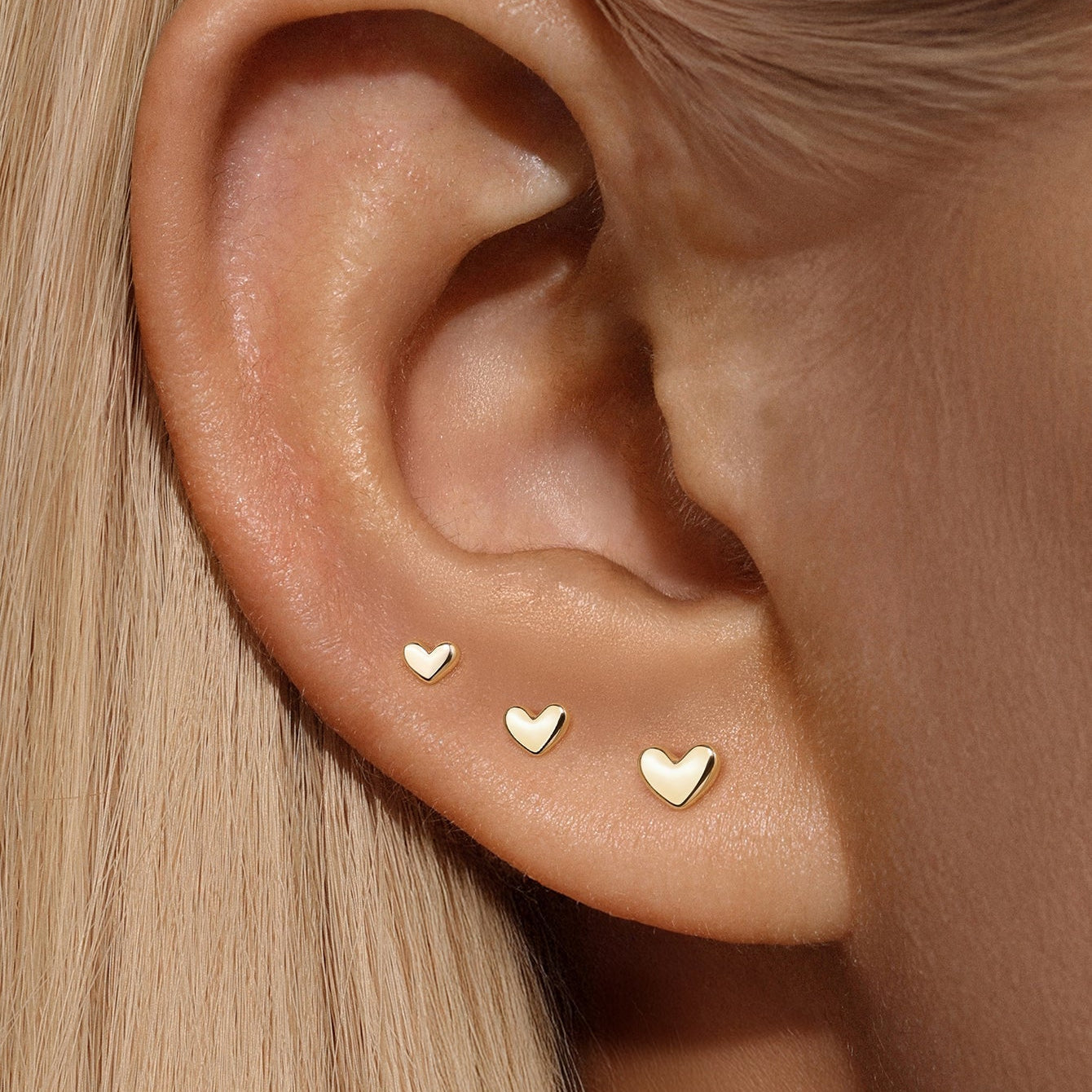 Model wearing three 14k gold heart studs on earlobe comparing small, medium, and large sizes for stacking.