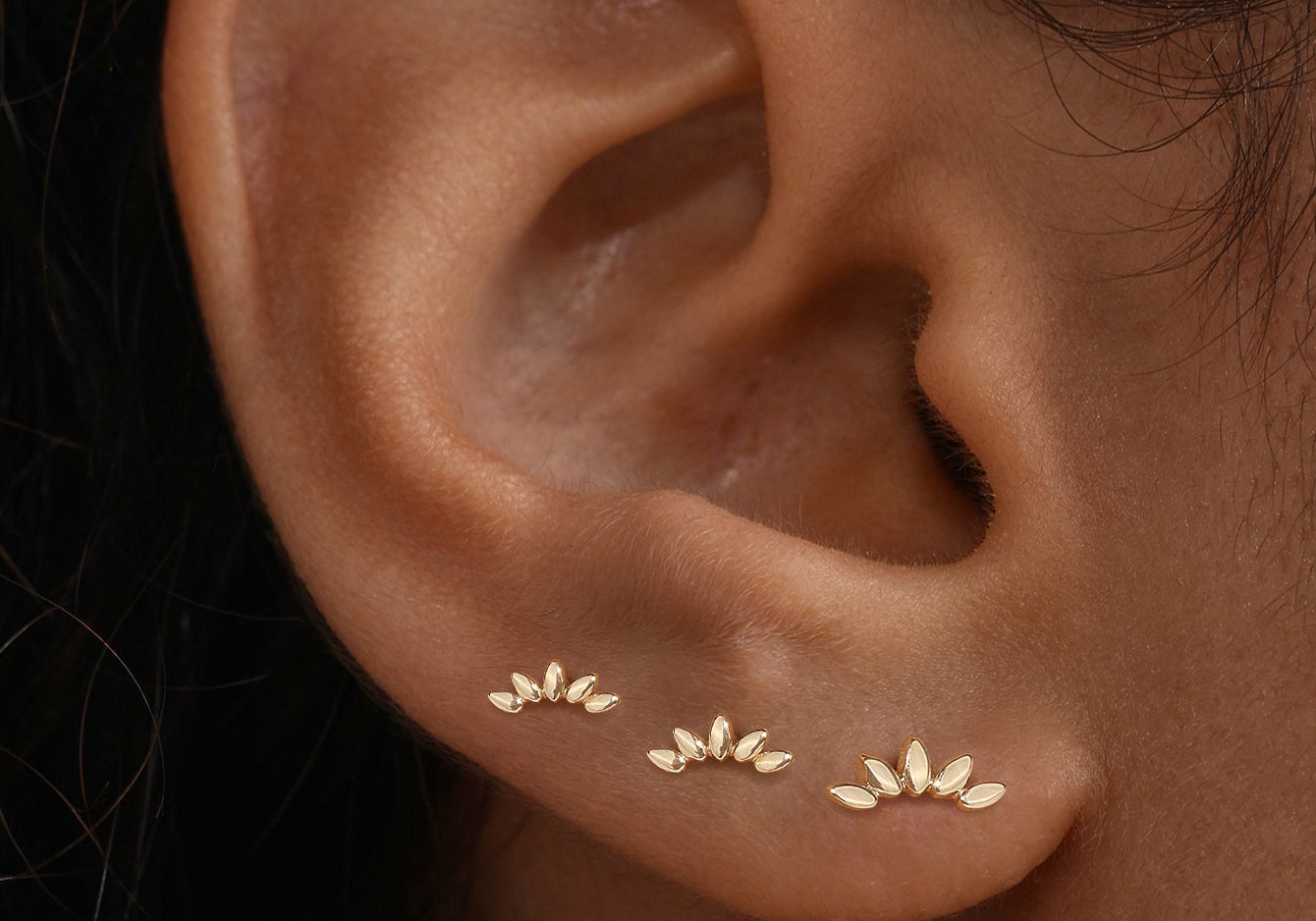 Three gold leaf-shaped stud earrings worn in a row along a person's lobe for a stacked jewelry look.