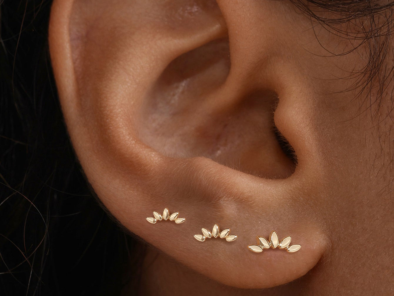 Three gold leaf-shaped stud earrings worn in a row along a person's lobe for a stacked jewelry look.