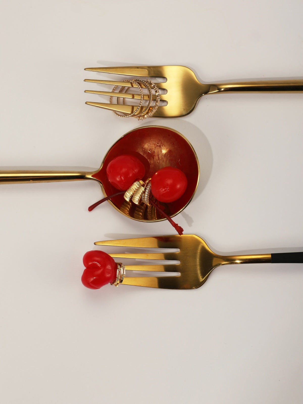 Stylized lifestyle shot of KOSINER ribbed striped mini hoop stacked with assorted gold cartilage jewelry, arranged on golden fork and spoon with red maraschino cherries on plain white background