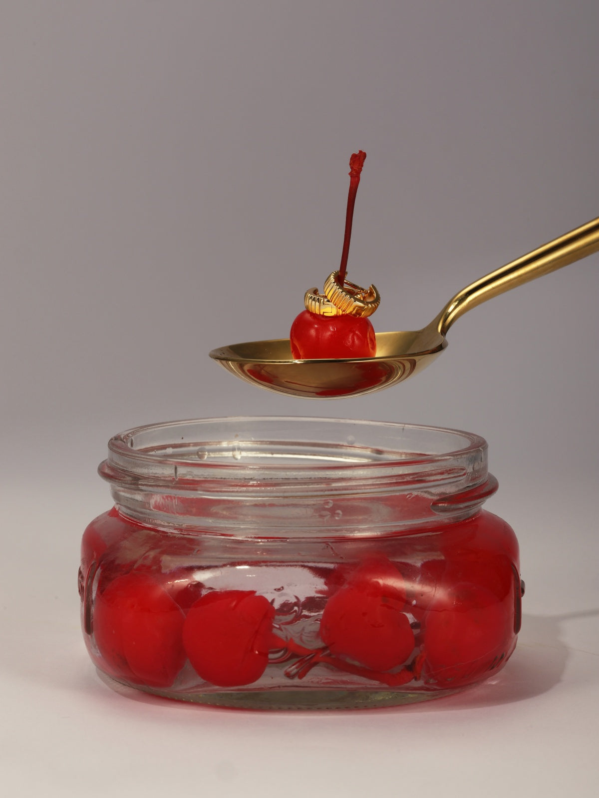 A single KOSINER horizontal striped ribbed hoop resting on a red cherry held by golden spoon above a glass jar filled with preserved cherries, minimalist food-inspired jewelry styling