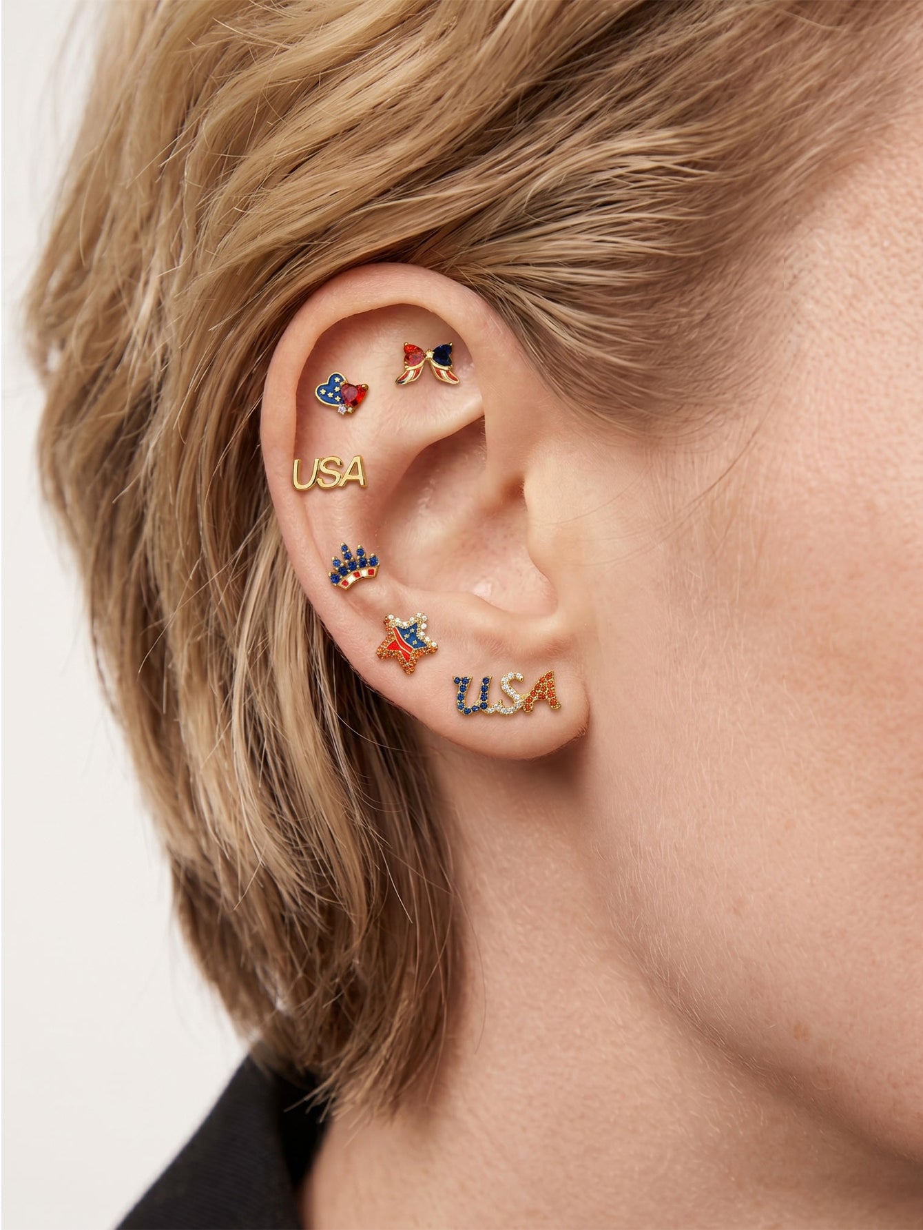 Patriotic USA themed ear stacking layout with KOSINER Independence Day patriotic USA letter flat back stud on earlobe, matched with heart, bow, crown and star flag-inspired cartilage piercings, festive multi-piercing styling for Fourth of July celebration