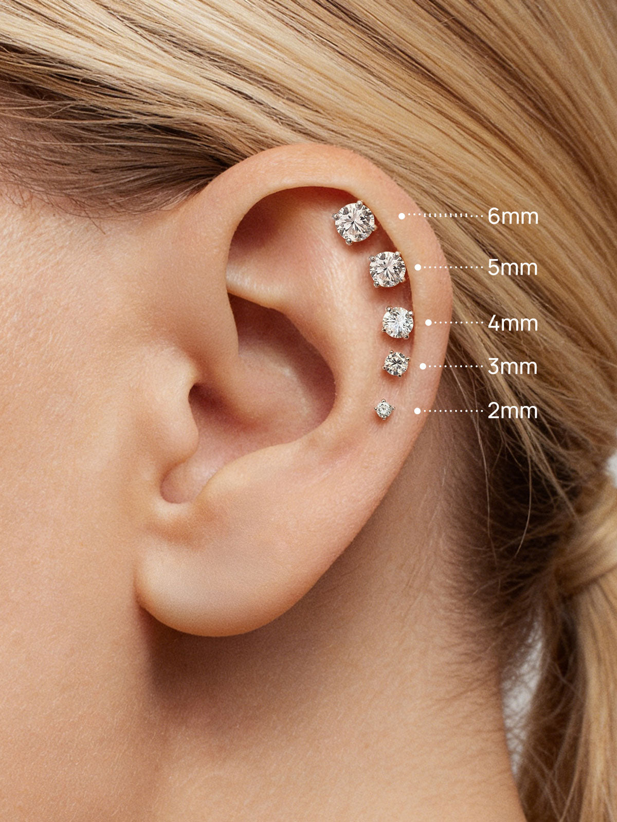 Close-up ear photo showing vertical helix stack of KOSINER lab-grown diamond flat back studs in sizes 2mm through 6mm, clearly labeled diameters for cartilage piercing size reference