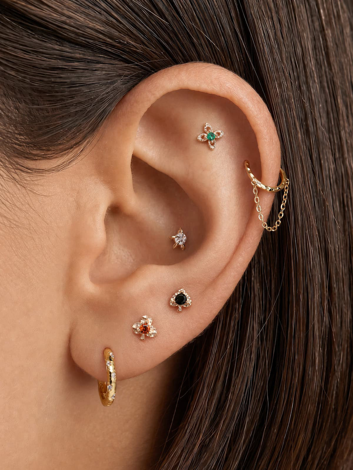 Stylish curated ear stack featuring KOSINER hand hammered black spade flat back stud on earlobe, paired with clover studs, four-petal flower helix stud, star conch stud and chain-linked ear cuff, poker-themed multi-piercing styling