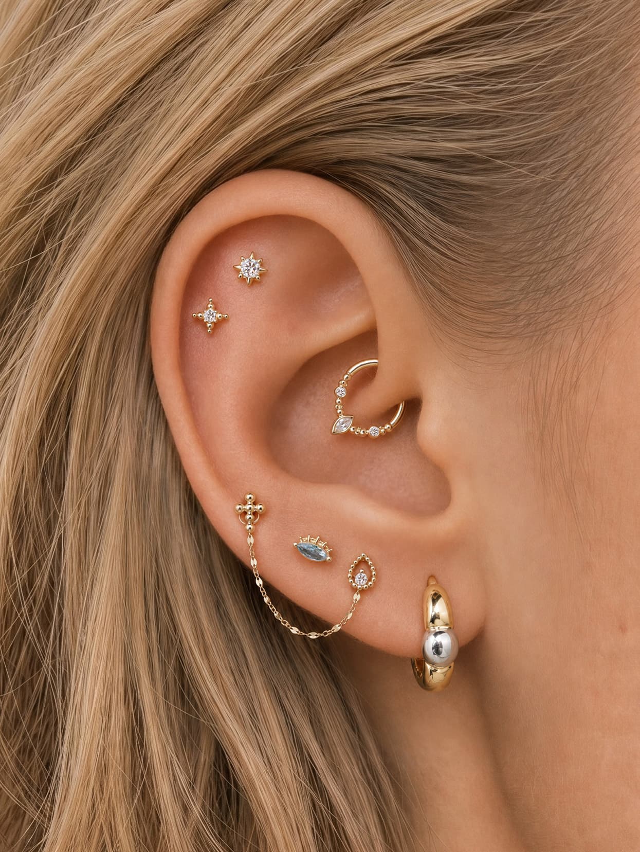 Elegant curated multi-piercing ear stack styled with KOSINER Perléa dainty star flat back helix studs, paired with mini cross stud, marquise daith hoop, chain-linked double lobe piercings, teardrop stud and pearl detail huggie hoop for sophisticated layered ear styling