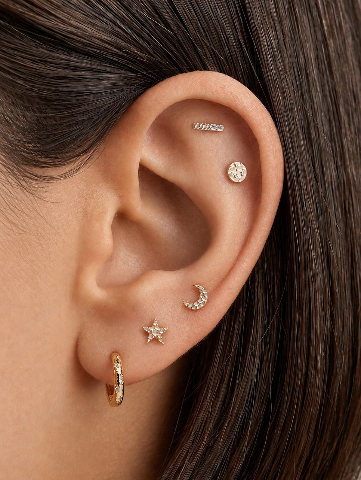 Curated ear stacking look with KOSINER hand hammered textured disc flat back stud on upper helix, matched with hammered star, moon, bar studs and textured huggie hoop, cohesive organic hammer-finish multi-piercing styling
