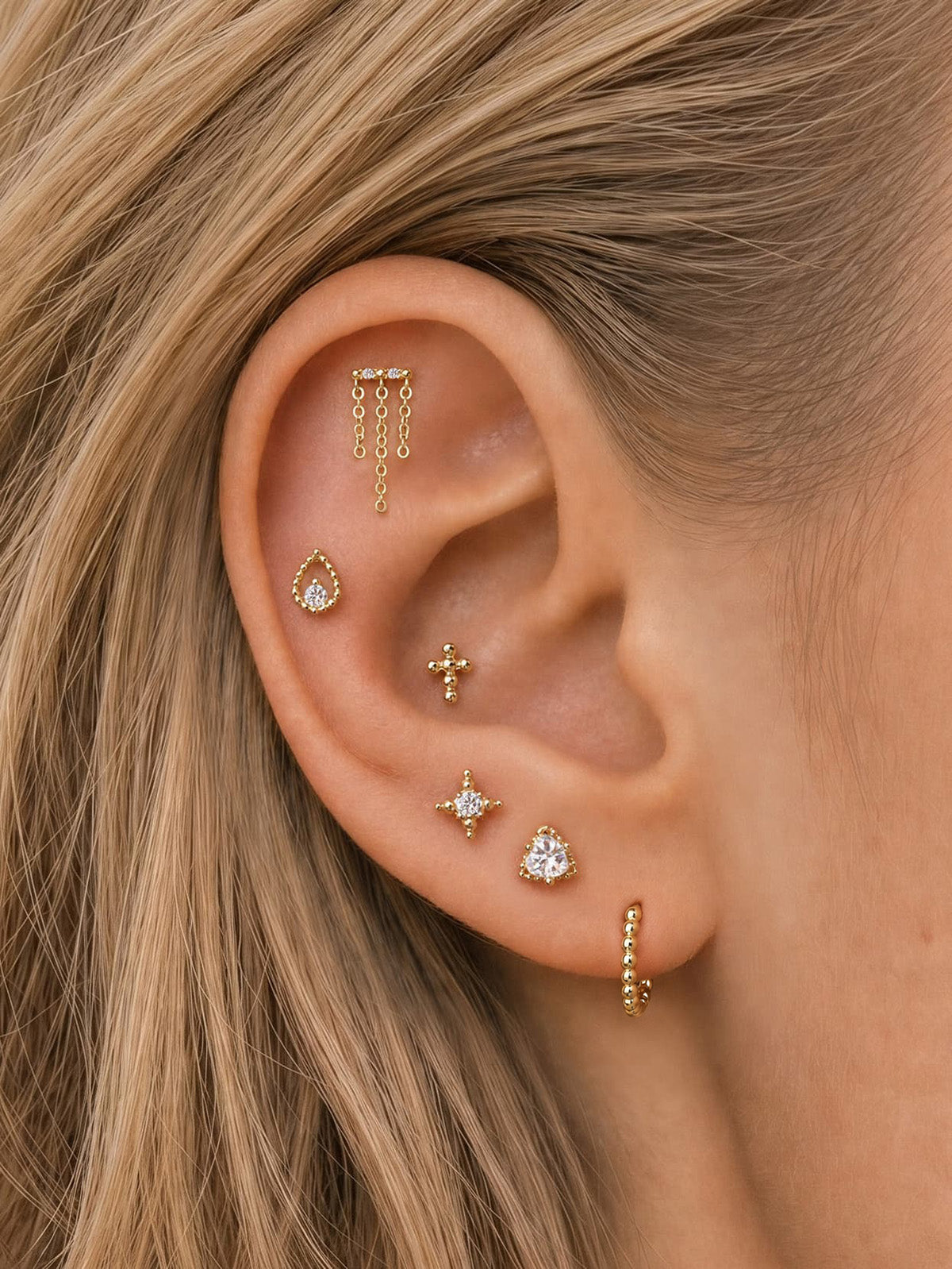 Elegant curated multi-piercing ear stack featuring KOSINER Perléa plain beaded hoop on earlobe, matched with chain drop helix stud, teardrop outline piercing, beaded cross stud, four-point star stud and heart-cut CZ stud, trendy layered ear styling design