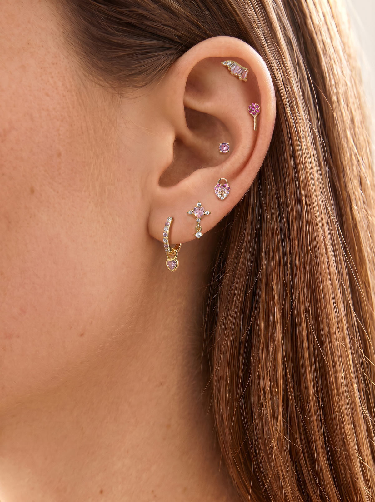 Romantic layered ear stacking look with KOSINER Pink Ritual heart lock flat back stud on earlobe, matched with cross drop, curved wing and round gem piercings, delicate multi-piercing daily styling