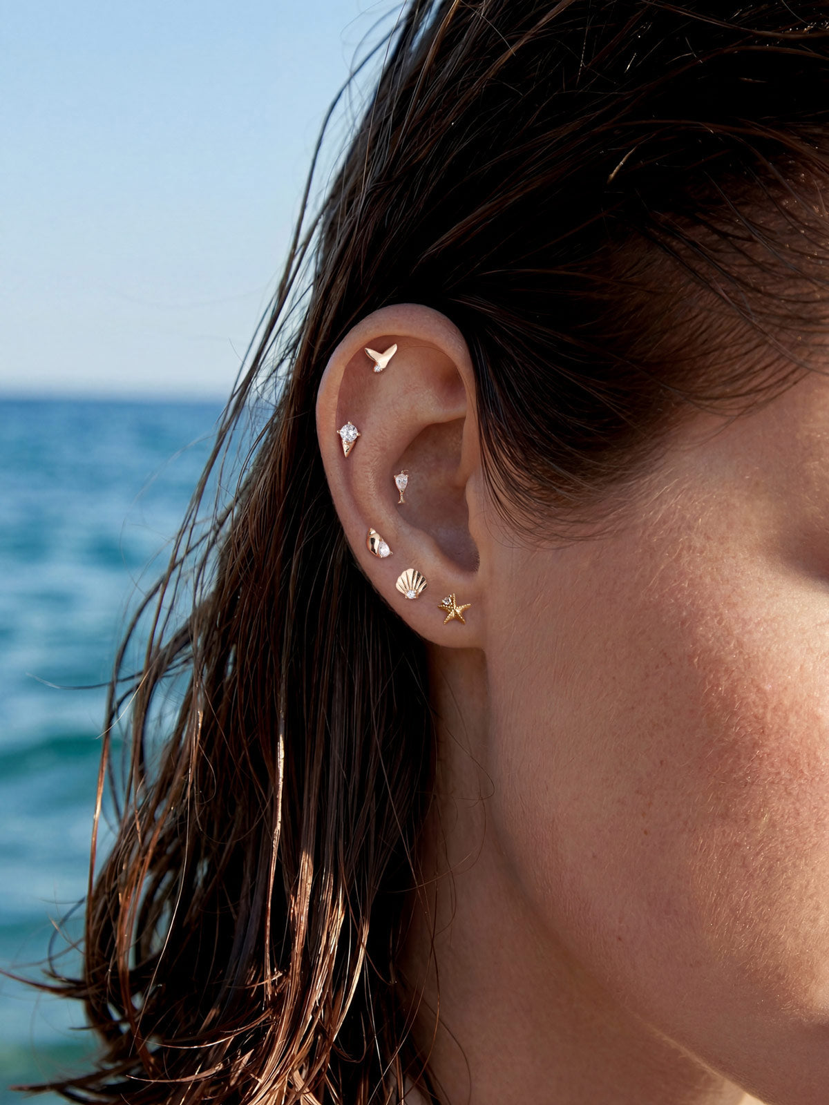 Trendy coastal full ear stacking design featuring KOSINER Seaspritz 14K solid gold seashell flat back stud, matched seastar, mermaid tail, cocktail glass and other ocean series tiny piercing studs, seaside multi-hole ear styling with ocean backdrop