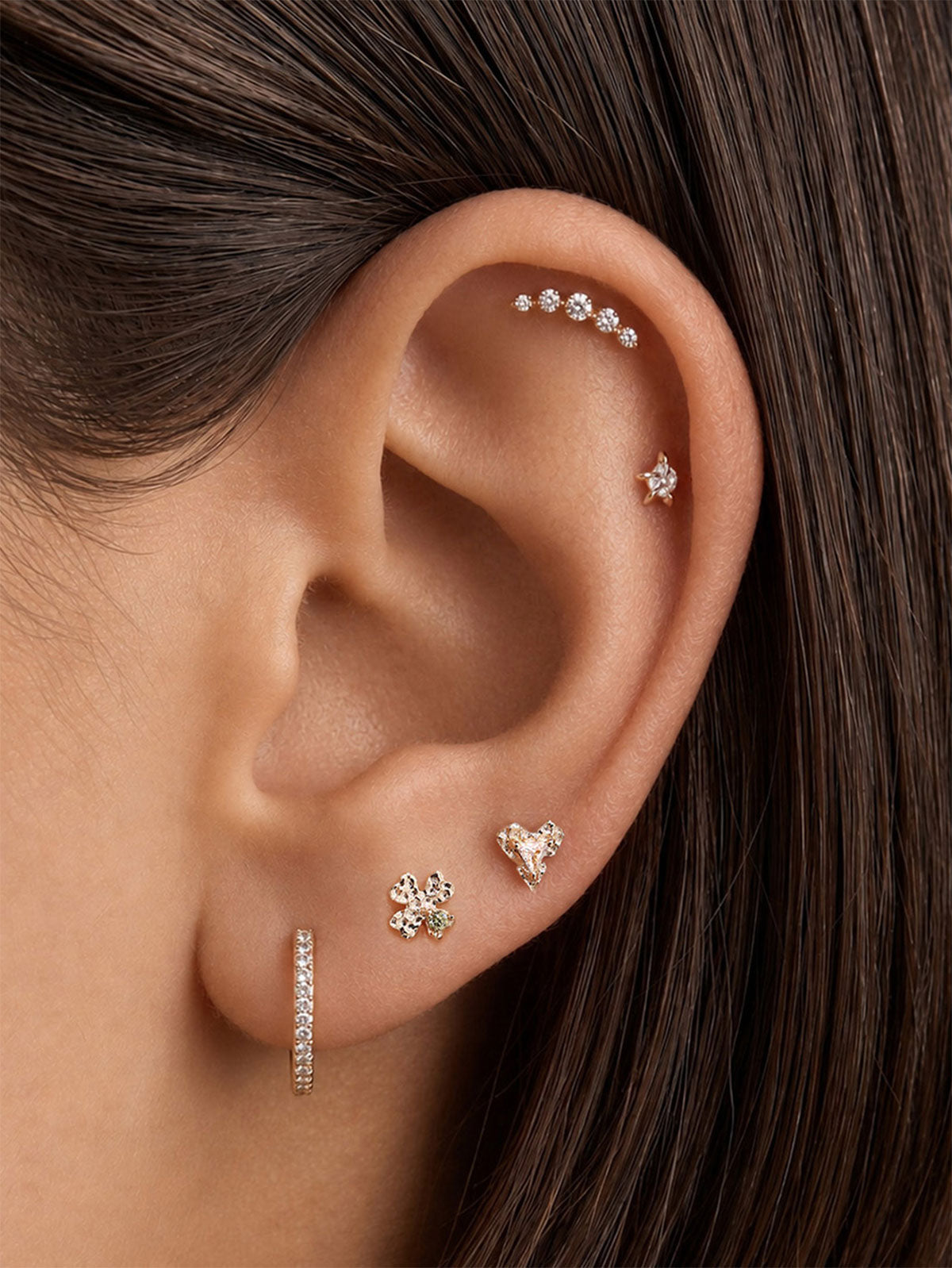 Curated multi-piercing ear styling with KOSINER hand hammered textured heart flat back stud worn on earlobe, matched with four-leaf clover stud, tiny star helix stud, linear CZ bar piercing and pavé huggie hoop, delicate layered ear stack layout