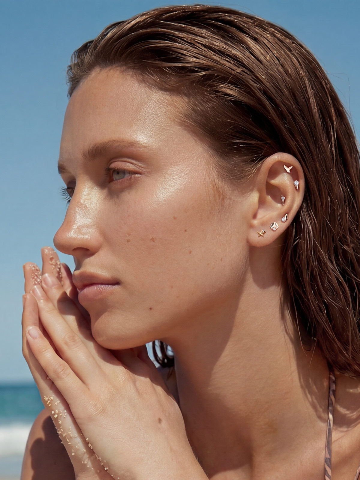 Beach lifestyle portrait of a model wearing KOSINER Seaspritz ocean themed piercing collection including textured seashell flat back stud, sun-kissed damp hair and sandy palms, bright seaside scenery for summer vacation jewelry inspiration