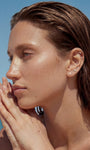 Beach lifestyle portrait of a model wearing KOSINER Seaspritz ocean themed piercing collection including polished dainty seahorse flat back stud, sun-kissed damp hair and sandy palms, bright seaside scenery for summer vacation jewelry inspiration