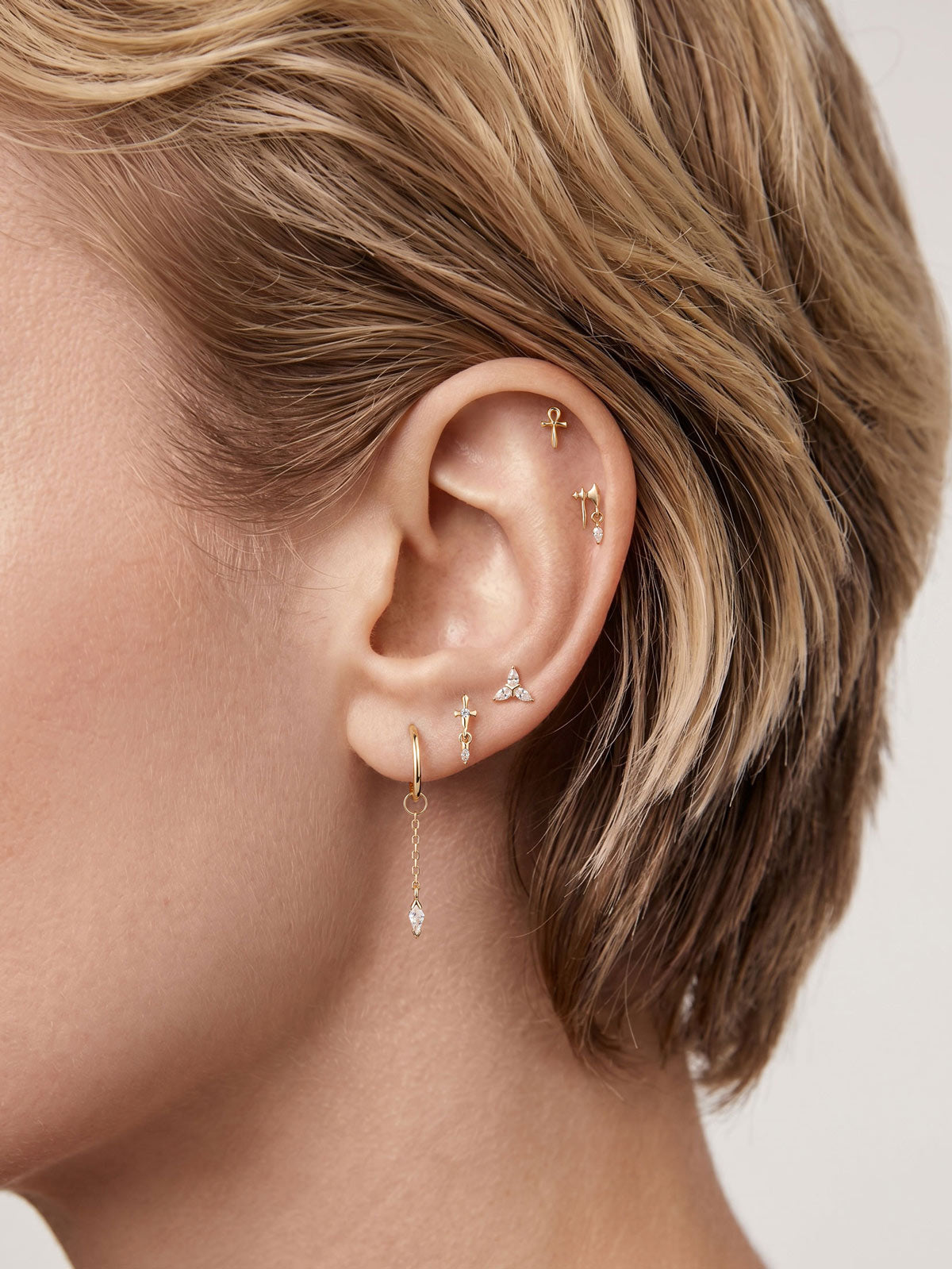 Short blonde hair model ear styling with 14K solid gold Trinity Knot tragus stud, assorted tiny gold piercing earrings close-up