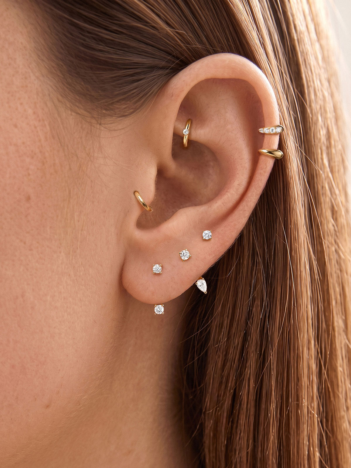 Close-up women's ear curated ear stack styling with KOSINER two-stone curved barbell helix piercing mixed with assorted gold hoop & stud earrings