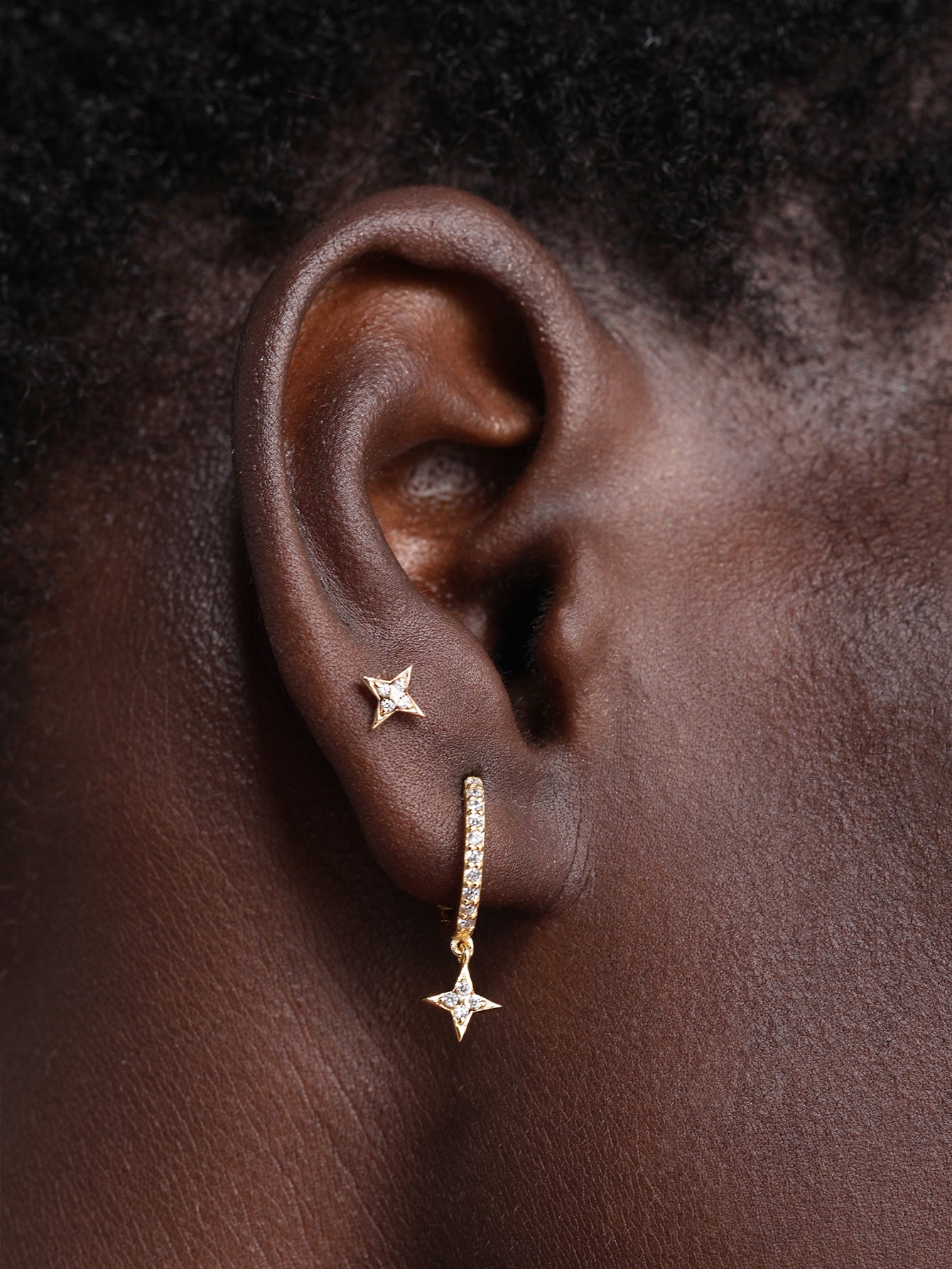 Close-up ear shot showing KOSINER Starlight dainty four-point star flat back stud worn on earlobe, paired with matching star charm hoop, demonstrating minimalist versatile fit on deep skin tone