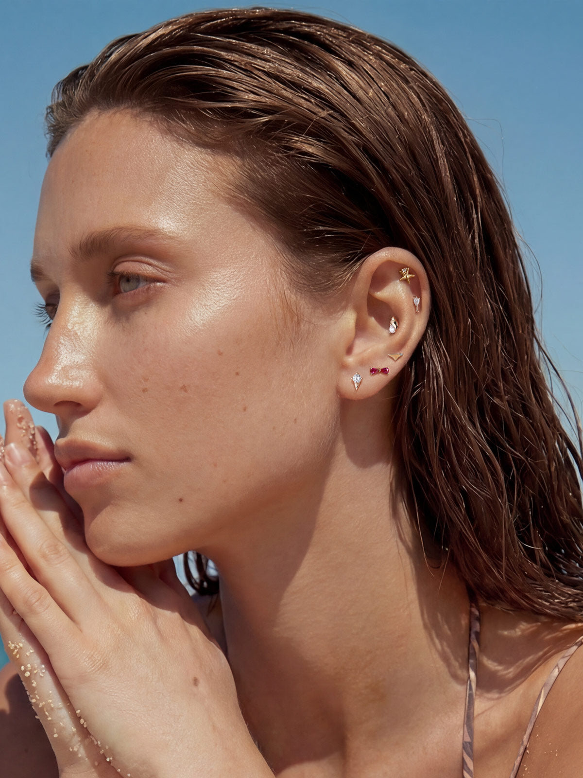 Beach lifestyle portrait of a model wearing the full KOSINER Seaspritz vacation collection including wine glass piercing stud, sun-wet hair and sandy palms, clear blue seaside background for holiday jewelry inspiration