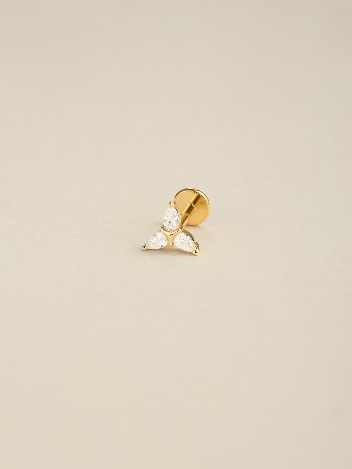 Angled still life photo of 14K solid gold Trinity Knot flat back labret stud, three pear shape cz ear piercing jewelry on soft beige background