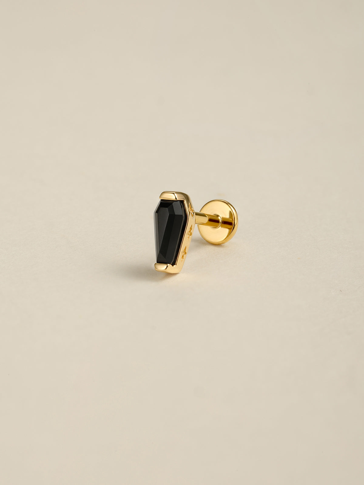 Angled still life photo of 14K solid gold black coffin flat back labret stud, black onyx goth ear piercing jewelry on soft beige background