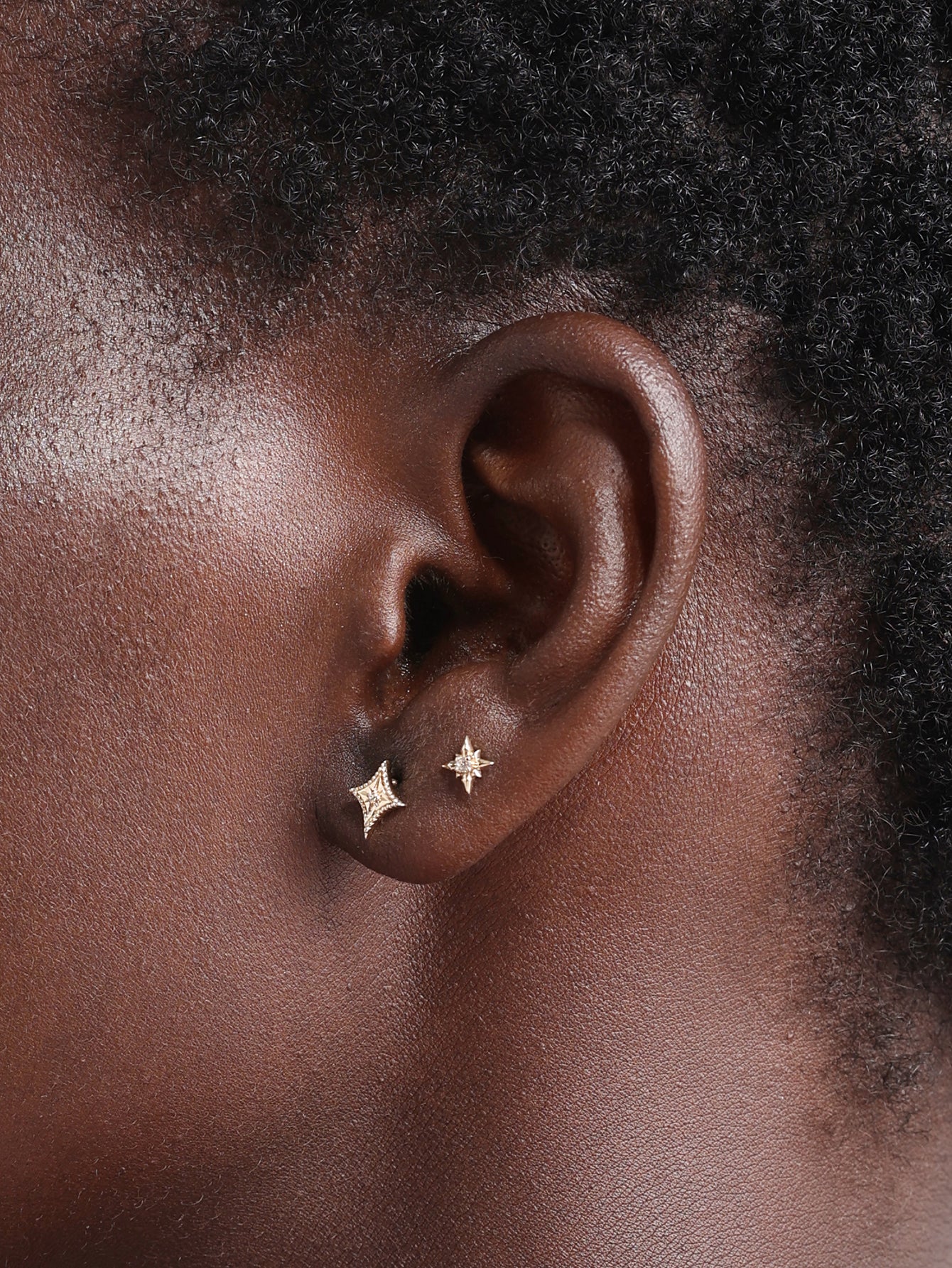 Close-up ear shot displaying two sizes of KOSINER Starlight four-point star flat back studs worn on lobe piercings, demonstrating minimalist versatile wear on deep complexioned skin