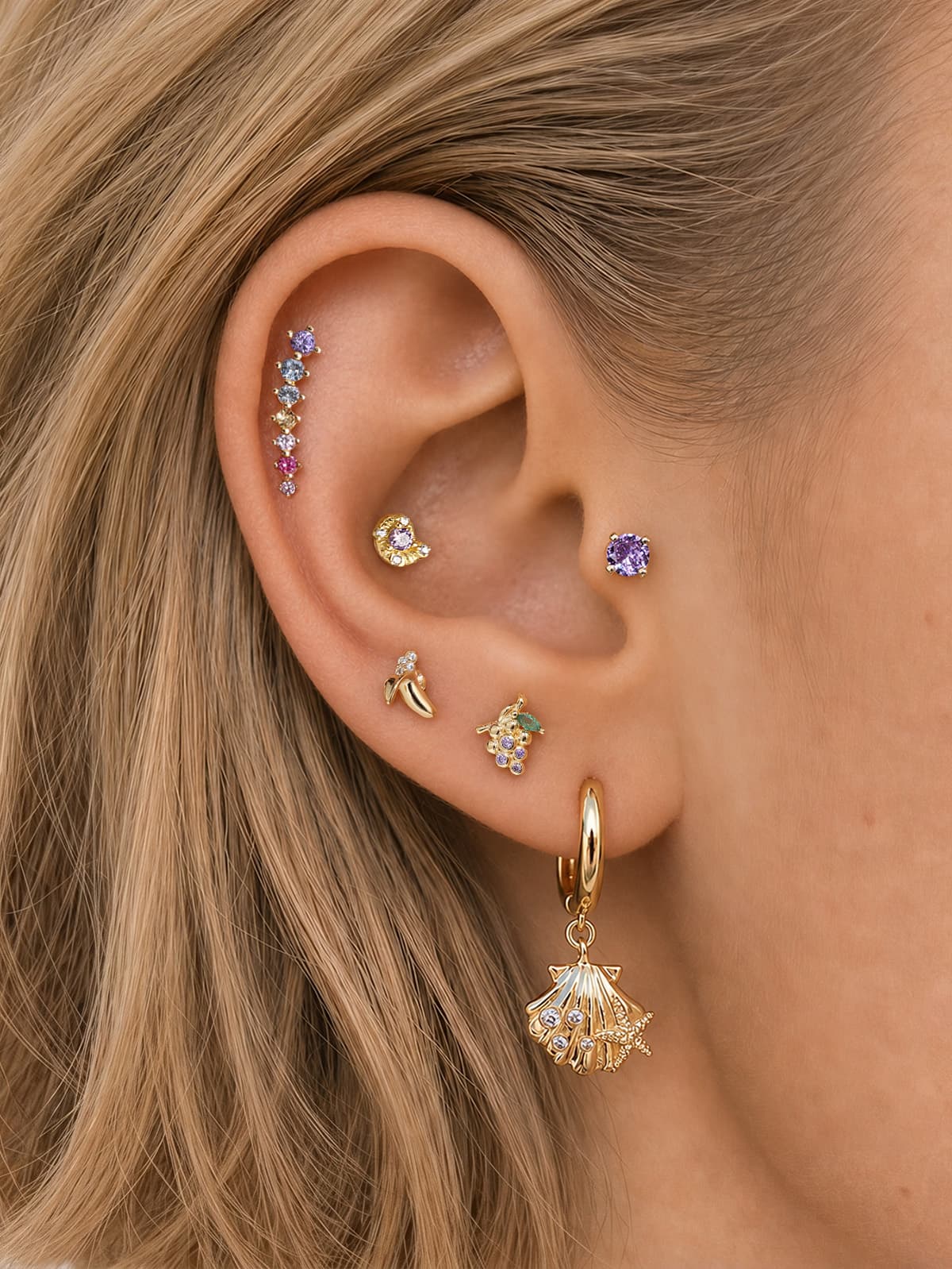 Mixed ear stack styling combining KOSINER fruit & sea jewelry lines, rainbow ear climber paired with banana, grape, seashell hoop, single purple gem piercings