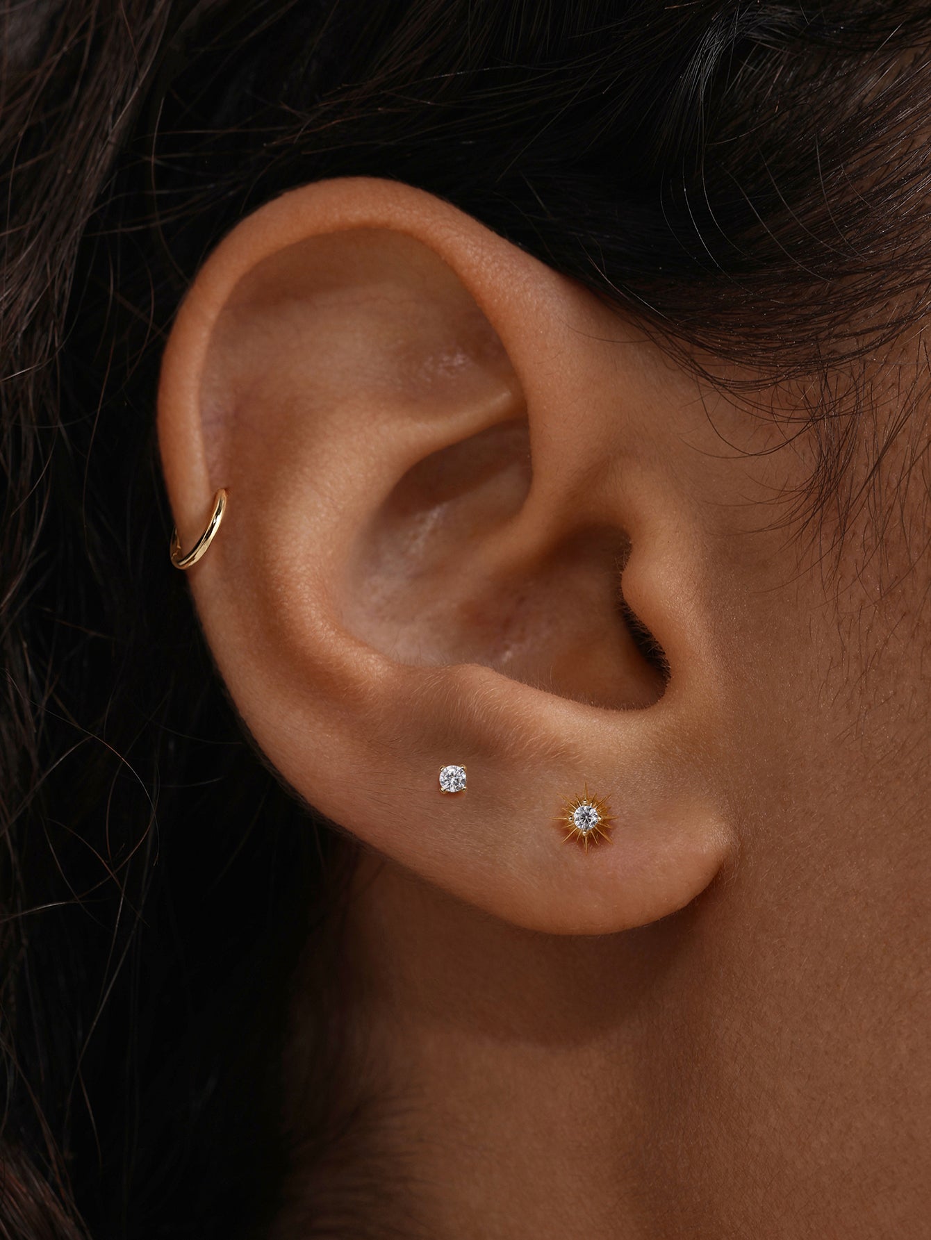 Close-up ear shot with KOSINER Étoile sunburst CZ flat back stud worn on earlobe, matched with simple solitaire stud and thin gold hoop, minimalist dainty everyday piercing combination