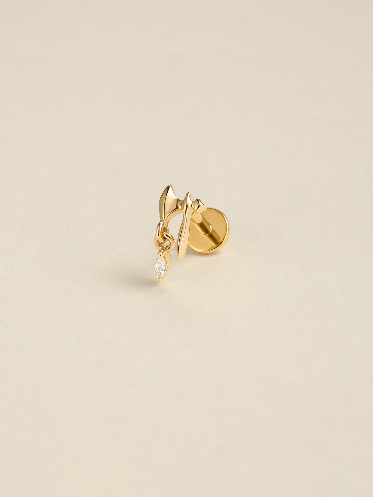 Angled still life shot of 14K solid gold battle axe drop flat back labret stud, clear marquise CZ dangle piercing jewelry on soft beige background