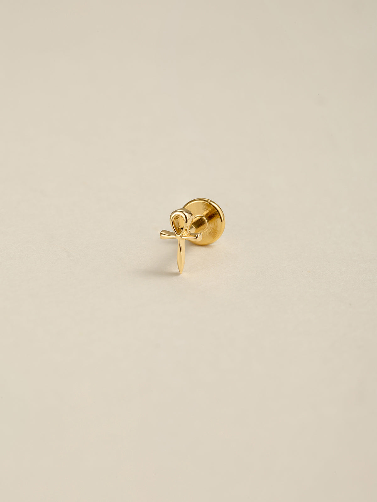 Angled still photograph of 14K solid gold Ankh cross flat back labret stud, Egyptian Ankh symbol earring on soft beige background