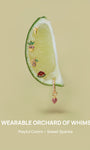 KOSINER Orchard whimsical wearable orchard jewelry laid on fresh lime slice, featuring watermelon, banana, pineapple, grape flat studs and strawberry charm clicker hoop, slogan A WEARABLE ORCHARD OF WHIMSY