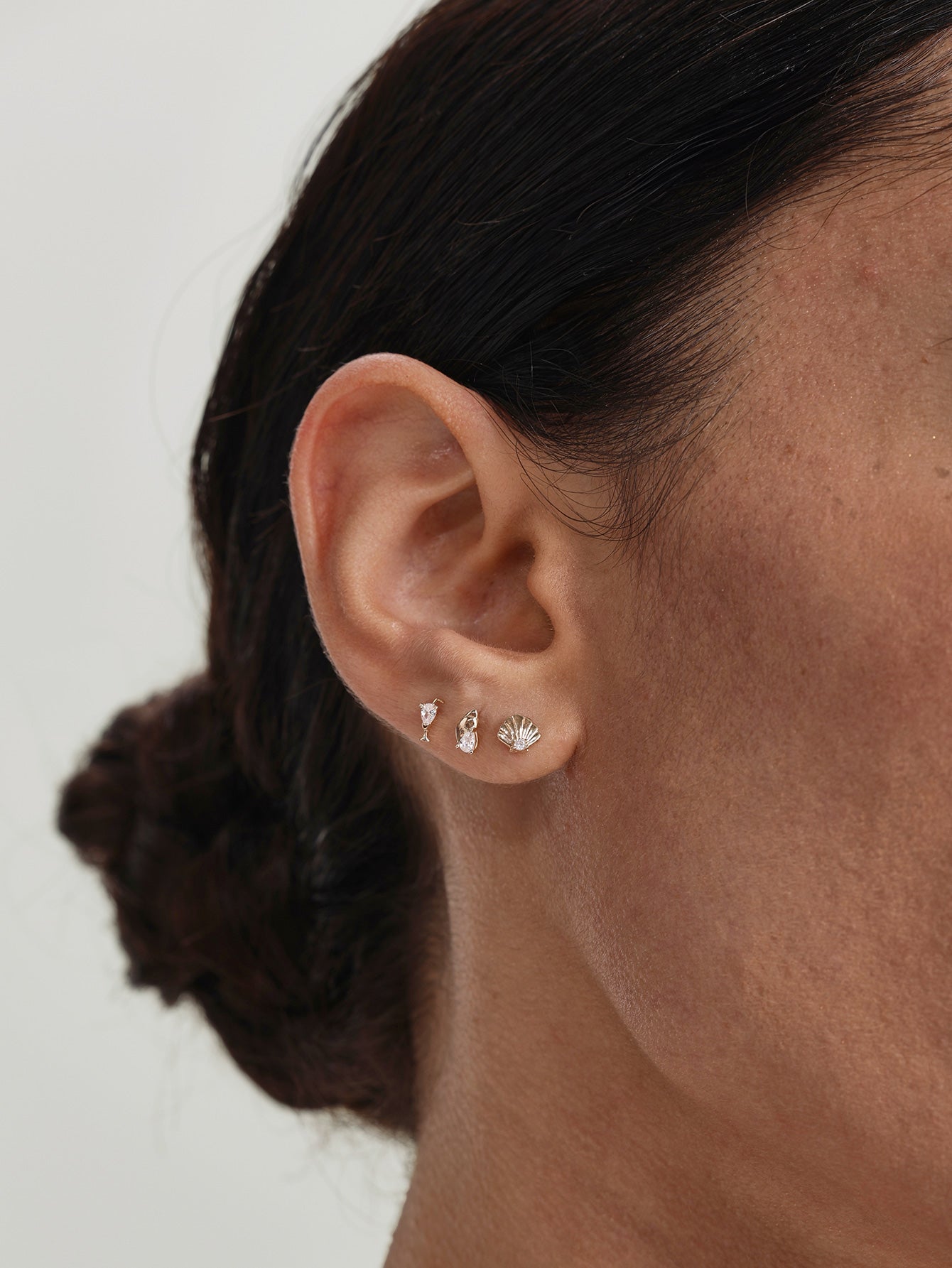 Close-up of human earlobe styled with KOSINER Seaspritz wine glass flat back stud, paired with seashell and other ocean-themed tiny studs, delicate minimalist daily ear stacking design