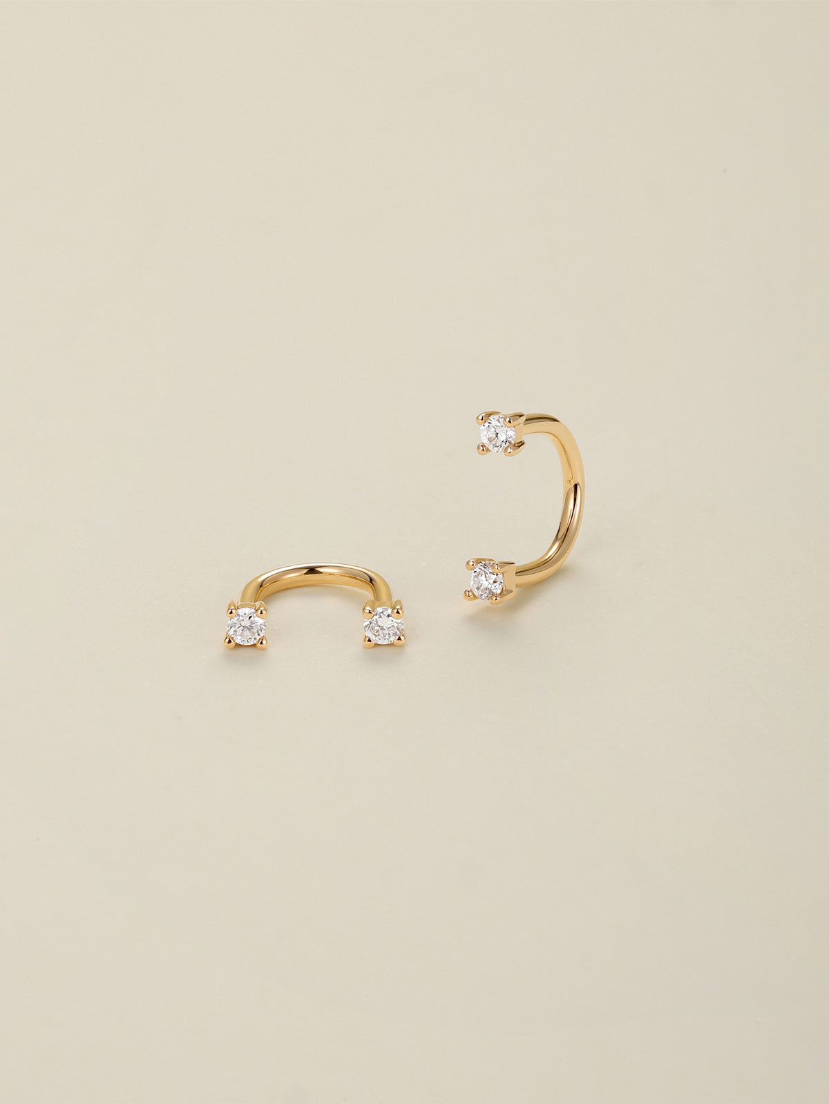 Pair of KOSINER 14K solid gold curved C barbell piercings, dual clear cubic zirconia gems secured by four-prong settings, laid flat on cream background