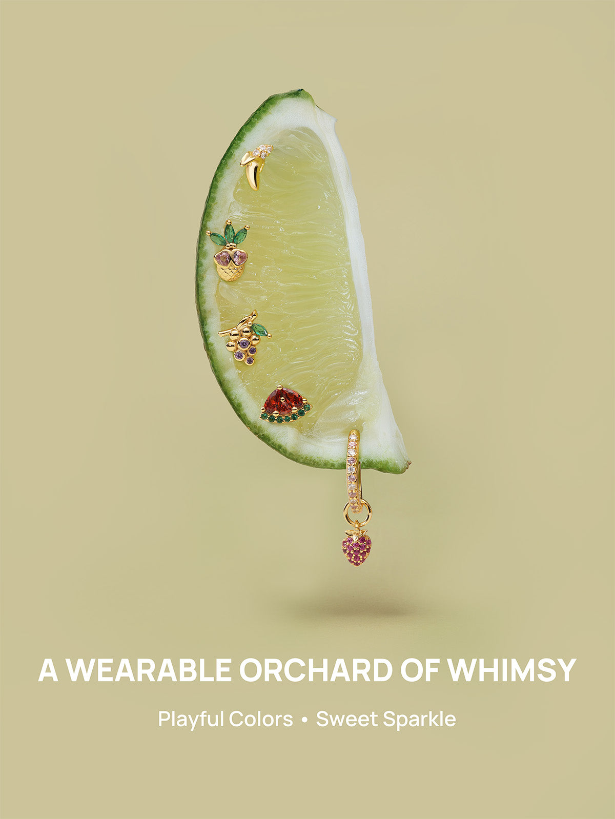 KOSINER Orchard whimsical fruit piercing jewelry laid on fresh lime slice, featuring pineapple, grape, watermelon, banana studs and strawberry dangle hoop, slogan A WEARABLE ORCHARD OF WHIMSY