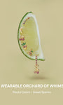 KOSINER Orchard wearable orchard jewelry laid on fresh lime slice, featuring cherry, banana, pineapple, grape studs and cherry dangle hoop, slogan A WEARABLE ORCHARD OF WHIMSY