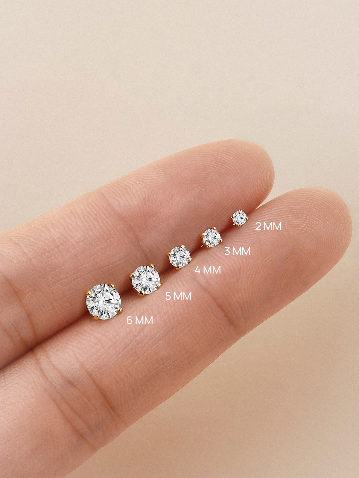 KOSINER lab-grown diamond flat back studs sized 2mm, 3mm, 4mm, 5mm, 6mm laid across fingertip to display scale difference of round brilliant gemstones in four-prong solid gold settings