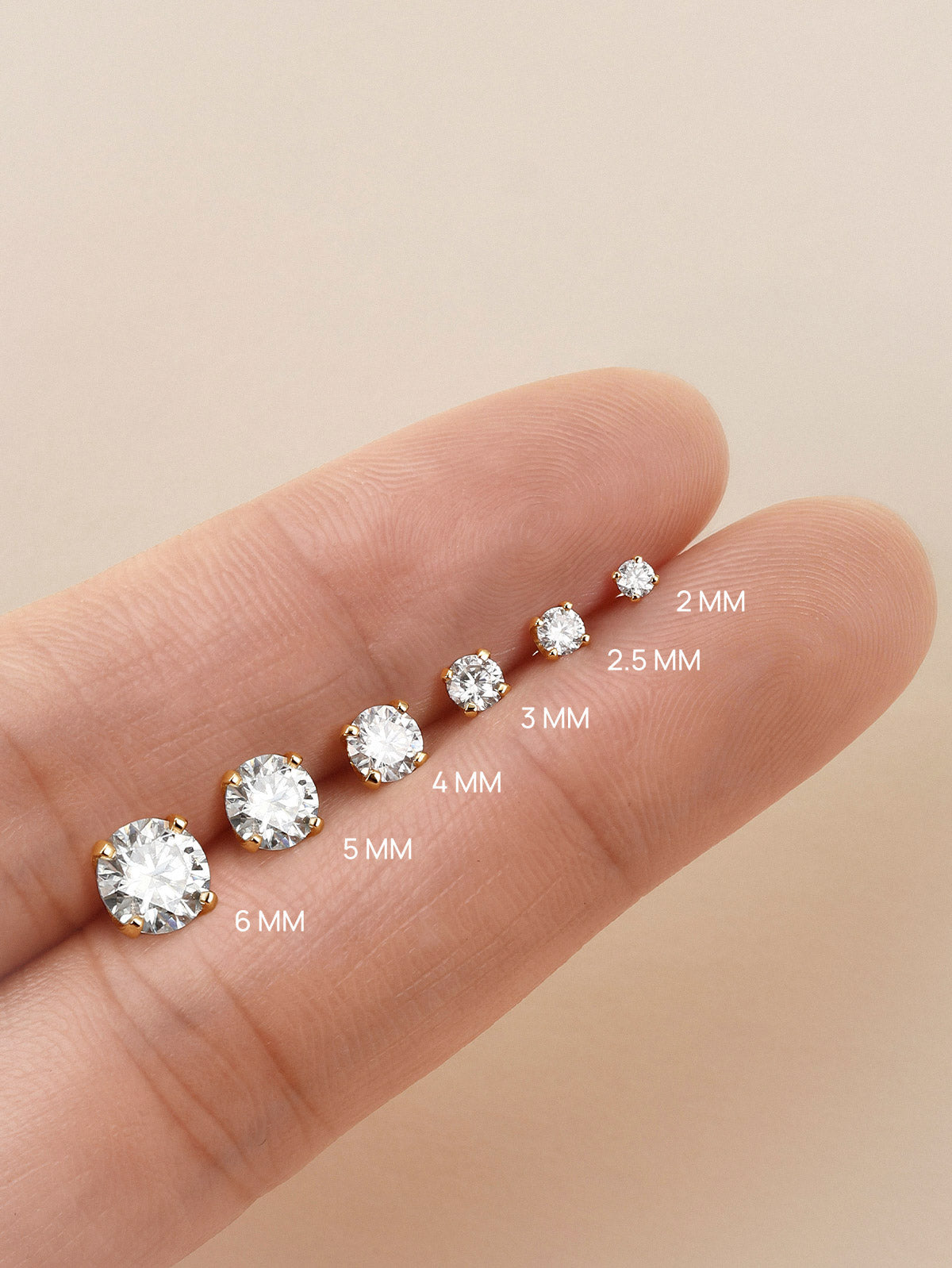 KOSINER moissanite flat back studs in 2mm to 6mm sizes laid on fingertip, showing visual scale difference of round brilliant-cut gemstones with four-prong gold settings
