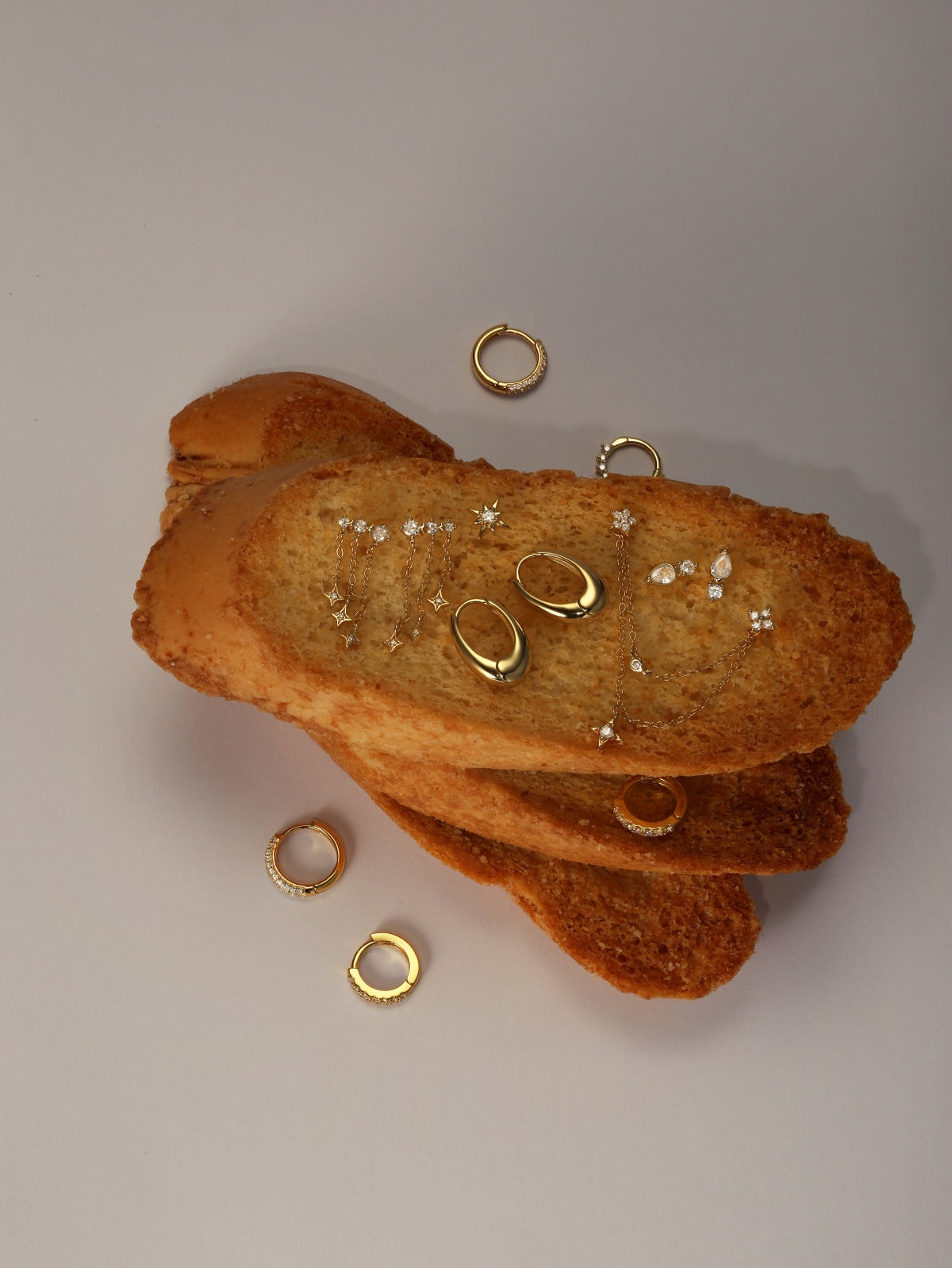 Aesthetic food-themed jewelry flat lay featuring KOSINER smooth oval drop gold hoops mixed with star chain dangling piercings and pavé crystal clicker hoops, arranged on stacked toast slices against neutral light gray background