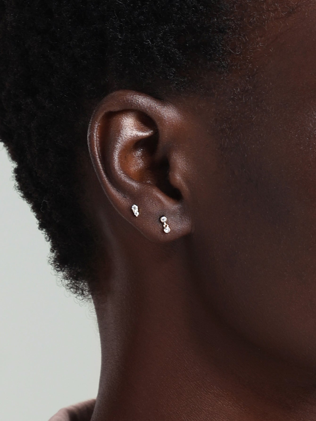 14K gold ear piercing studs with clear stones, one stacked vertically, one with a dangling bar, shown on ear.